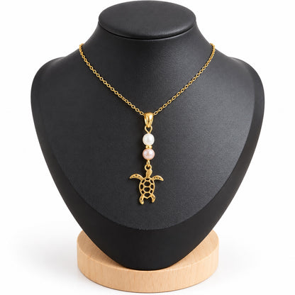 Collier tortue doré avec perles d’eau douce blanche et rosée sur présentoir noir, fond blanc