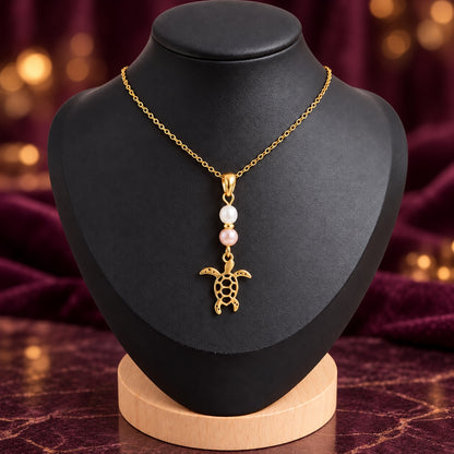 Collier tortue doré avec perles d’eau douce blanche et rosée sur présentoir noir, fond chic violet et doré